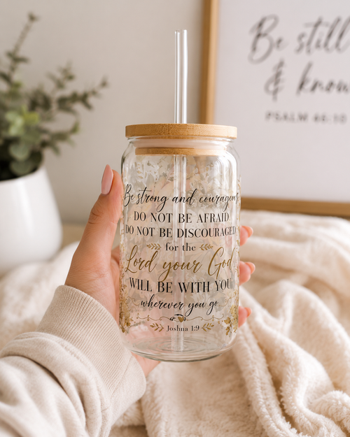 A hand holding a clear glass mason jar tumbler with a bamboo lid and a clear straw. The glass features a printed inspirational bible verse in black and gold lettering.