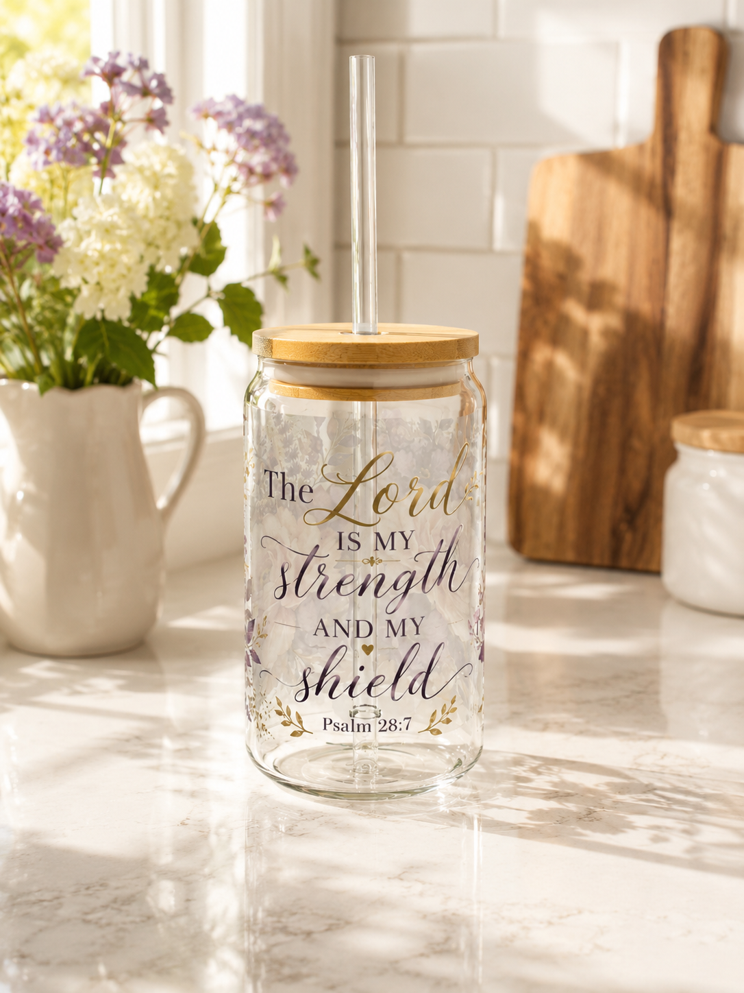 A clear glass mason jar tumbler with a bamboo lid and clear straw, featuring the printed text 'The Lord IS MY strength AND MY shield Psalm 28:7' in black and gold lettering, set on a marble countertop in a bright kitchen.