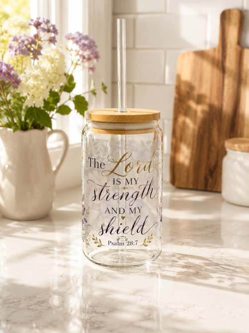 A clear glass mason jar tumbler with a bamboo lid and clear straw, featuring the printed text 'The Lord IS MY strength AND MY shield Psalm 28:7' in black and gold lettering, set on a marble countertop in a bright kitchen.