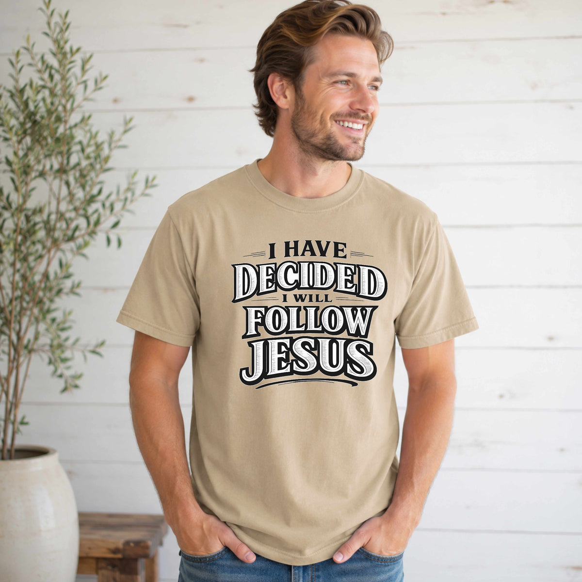 Man wearing a beige t-shirt with 'I Have Decided I Will Follow Jesus' text, standing against a light wooden paneled wall.