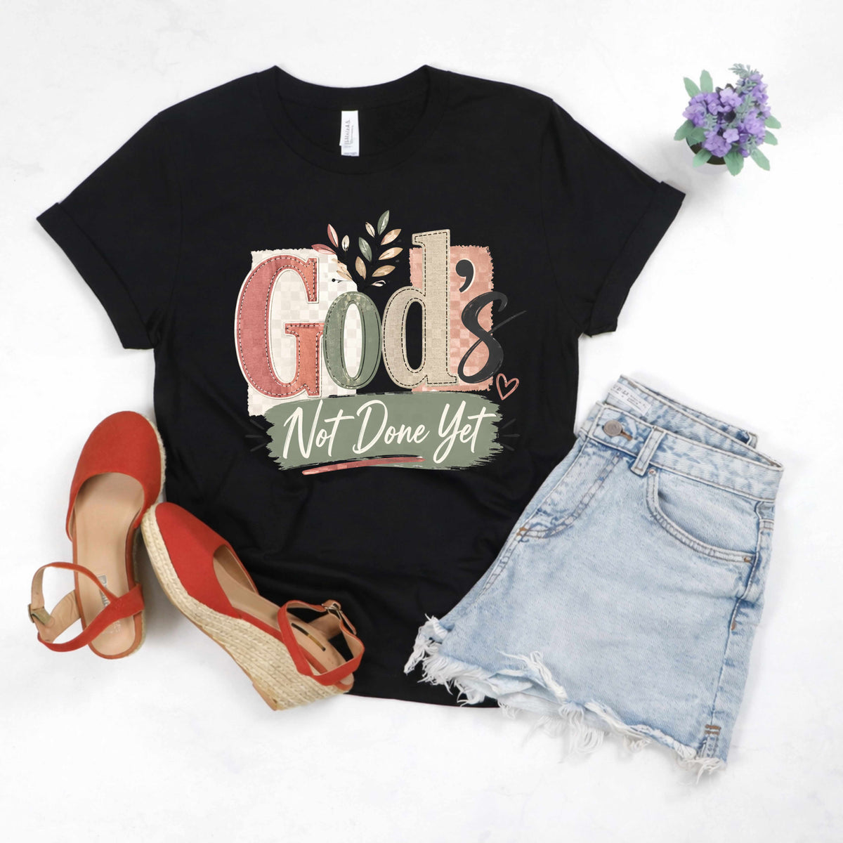 Black t-shirt with 'God's Not Done Yet' text, red shoes, denim shorts, and a small plant on a white background