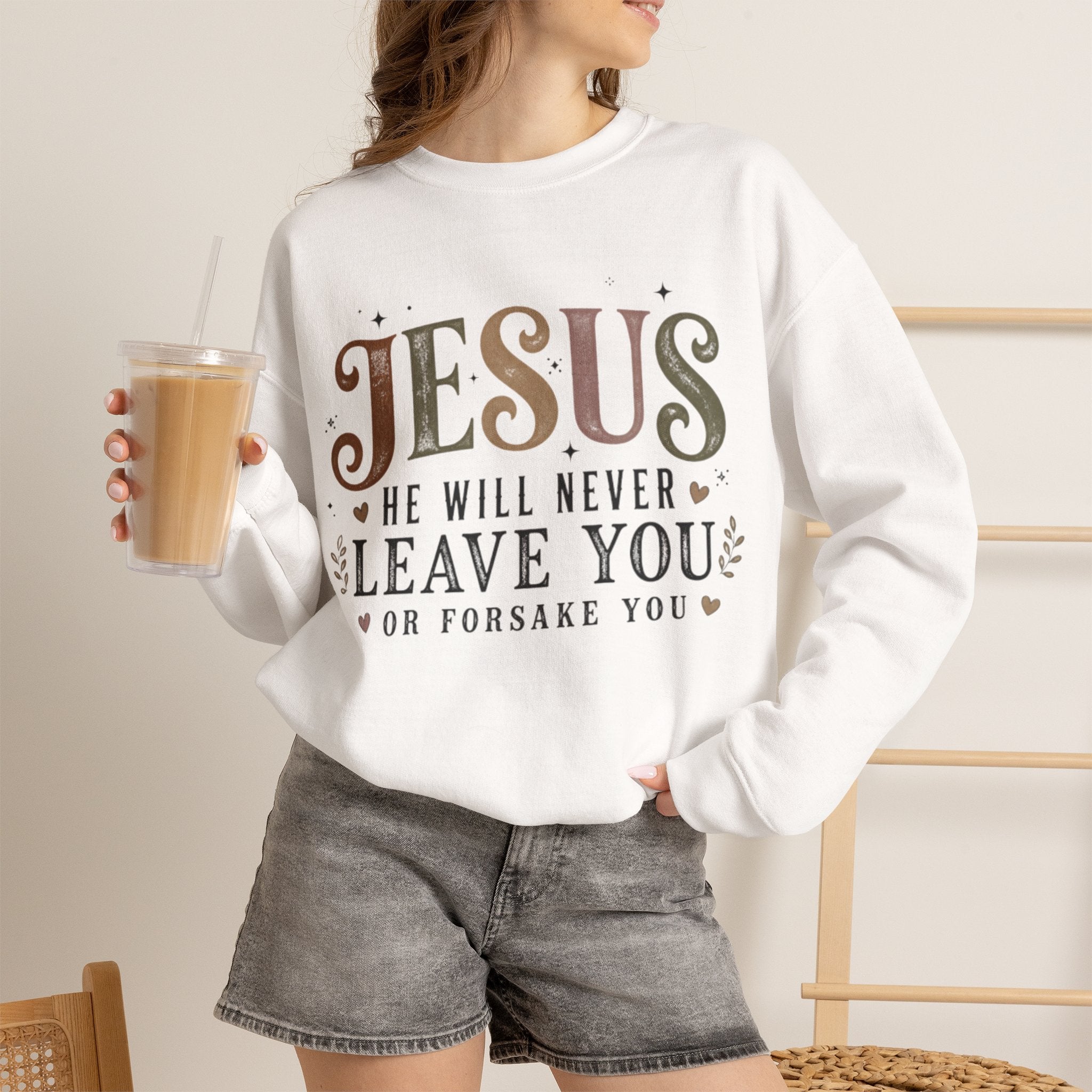 A woman wearing a white crewneck sweatshirt with a graphic that reads 'JESUS HE WILL NEVER LEAVE YOU OR FORSAKE YOU' in brown and black lettering. She is holding an iced coffee and wearing gray denim shorts in a warm, neutral indoor setting.