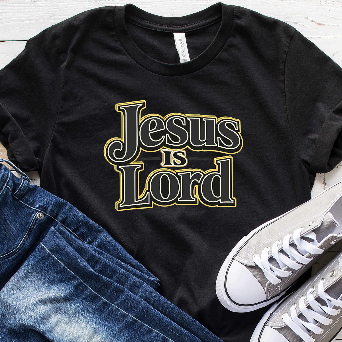 Black t-shirt with 'Jesus is Lord' text, paired with jeans and white sneakers on a wooden surface.