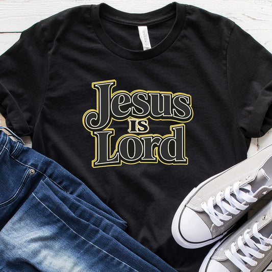 Black t-shirt with 'Jesus is Lord' text, paired with jeans and white sneakers on a wooden surface.