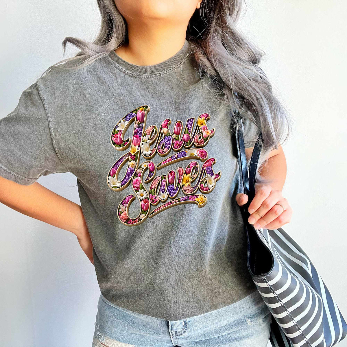 Person wearing a gray t-shirt with 'Jesus Saves' in colorful letters, holding a striped bag.