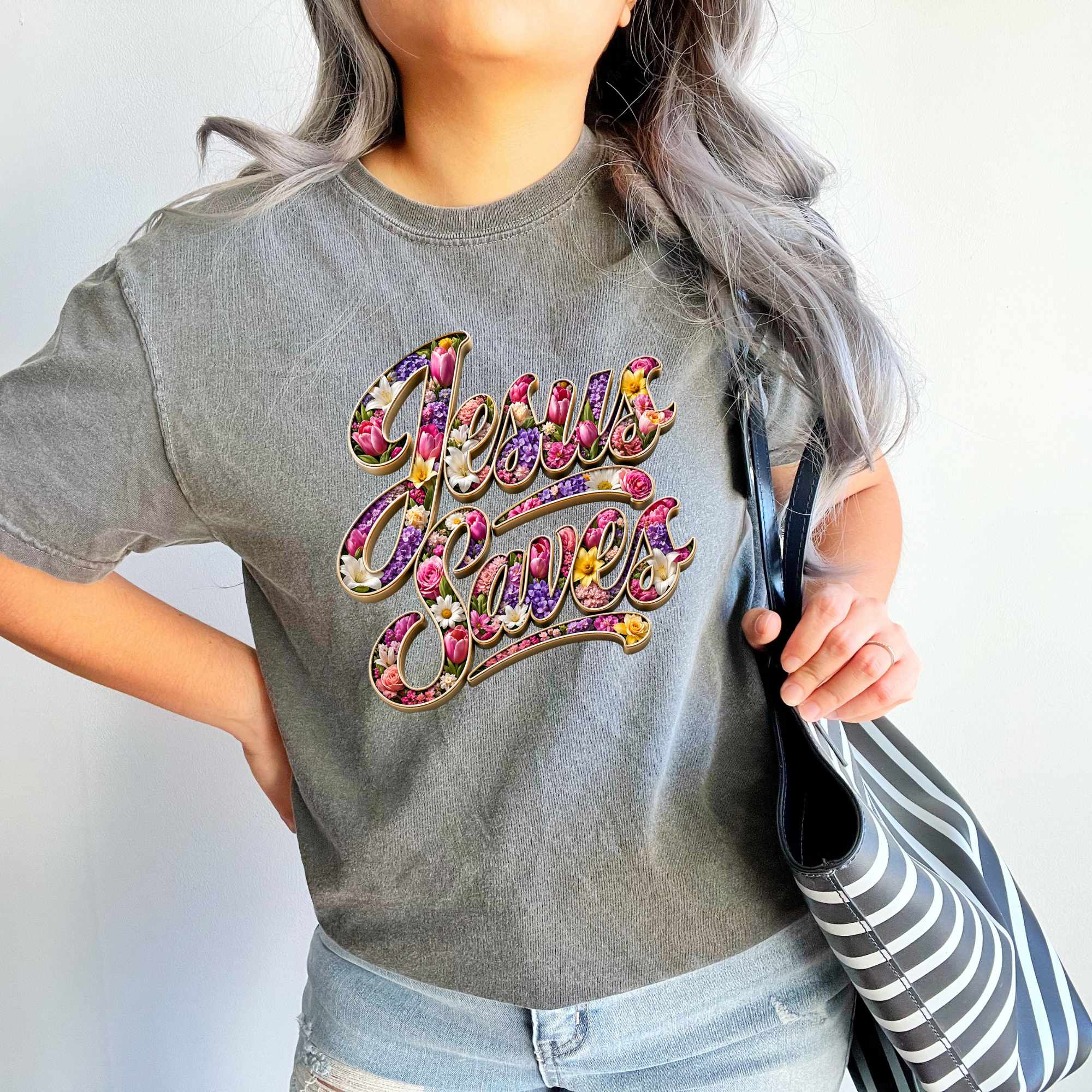 Person wearing a gray t-shirt with 'Jesus Saves' in colorful letters, holding a striped bag.
